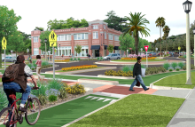 Color rendering of people walking or bicycling at an intersection with a roundabout, crosswalks, protected bike lanes, trees and grass, and a 2-story building in the background.